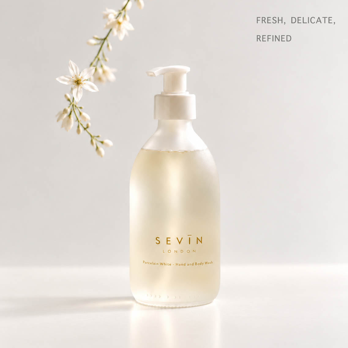 A frosted glass pump bottle of SEVIN LONDON Persaline Hair + Body Wash with pale yellow liquid inside, set against a clean white background with a delicate white flower sprig on the left and the text 'FRESH, DELICATE, REFINED' in the top right.