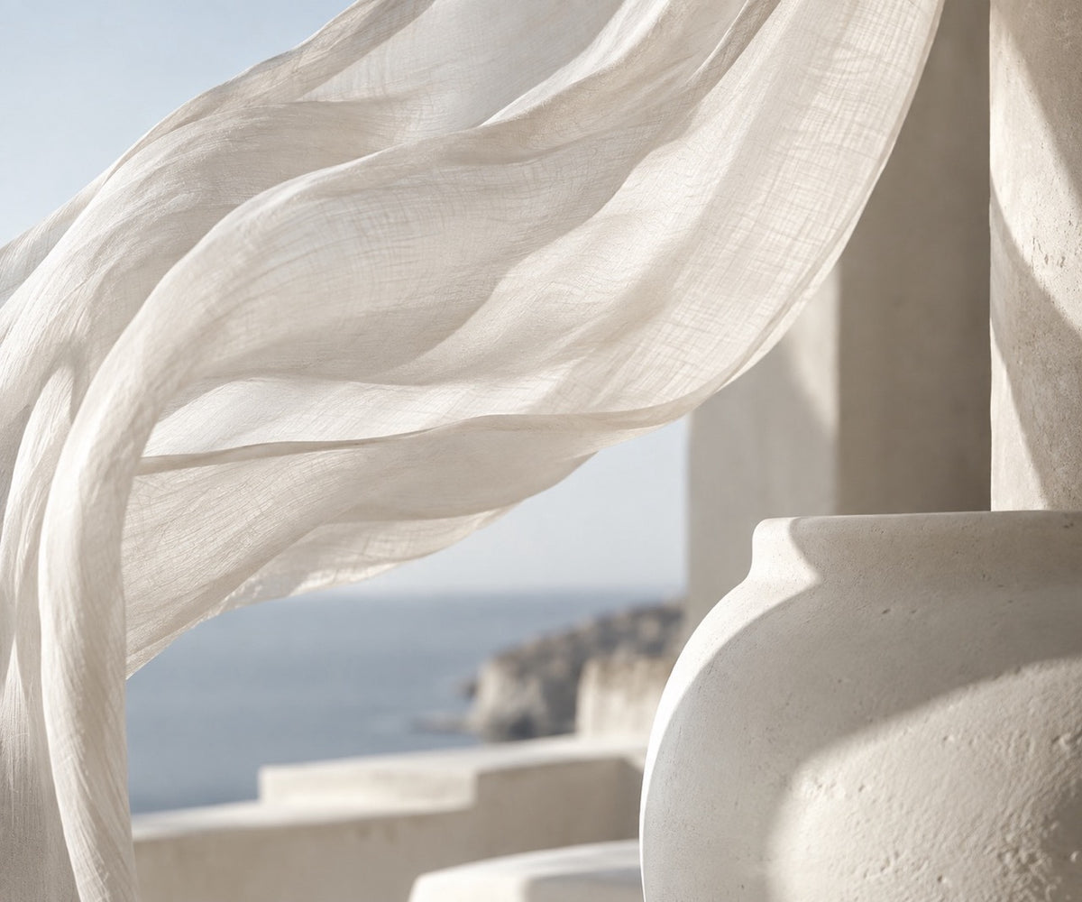 A flowing sheer white fabric drape in the wind beside a textured white ceramic planter, with a blurred coastal background.