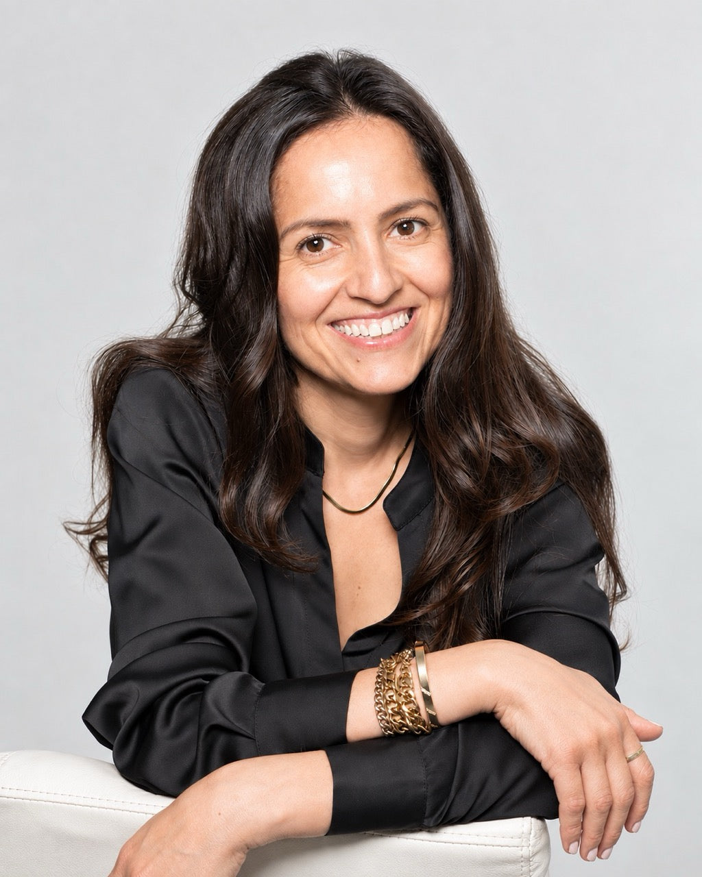 A smiling woman with long dark brown hair, wearing a black satin blouse and gold bracelets, leaning on a white chair against a light gray background. Picture of Sevin Ersin, Founder of Sevin London.