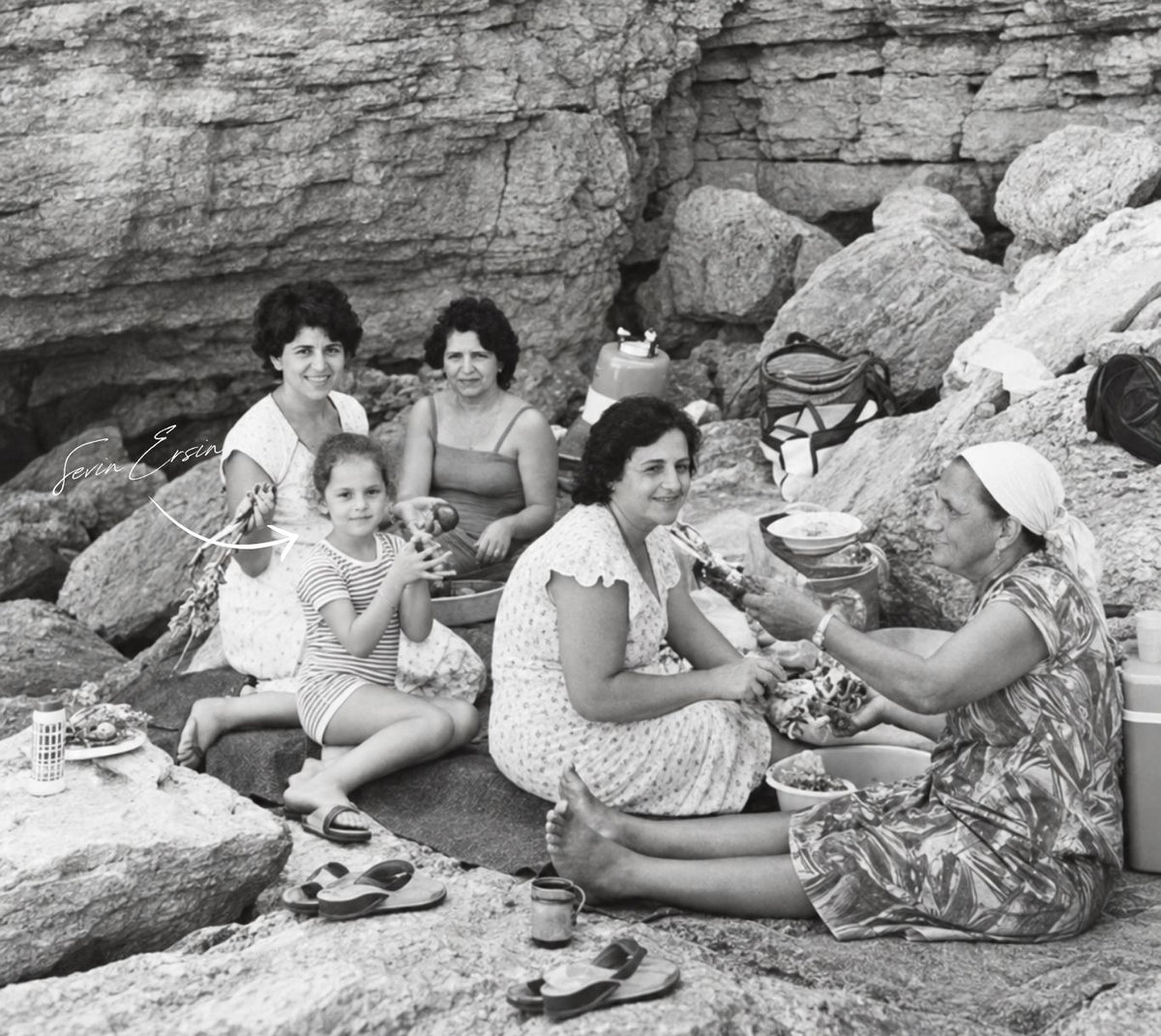 Black and white photograph of a group of five women and a child (Founder Sevin and her family) sitting together on rocks outdoors, sharing food and drinks in a rocky natural setting.