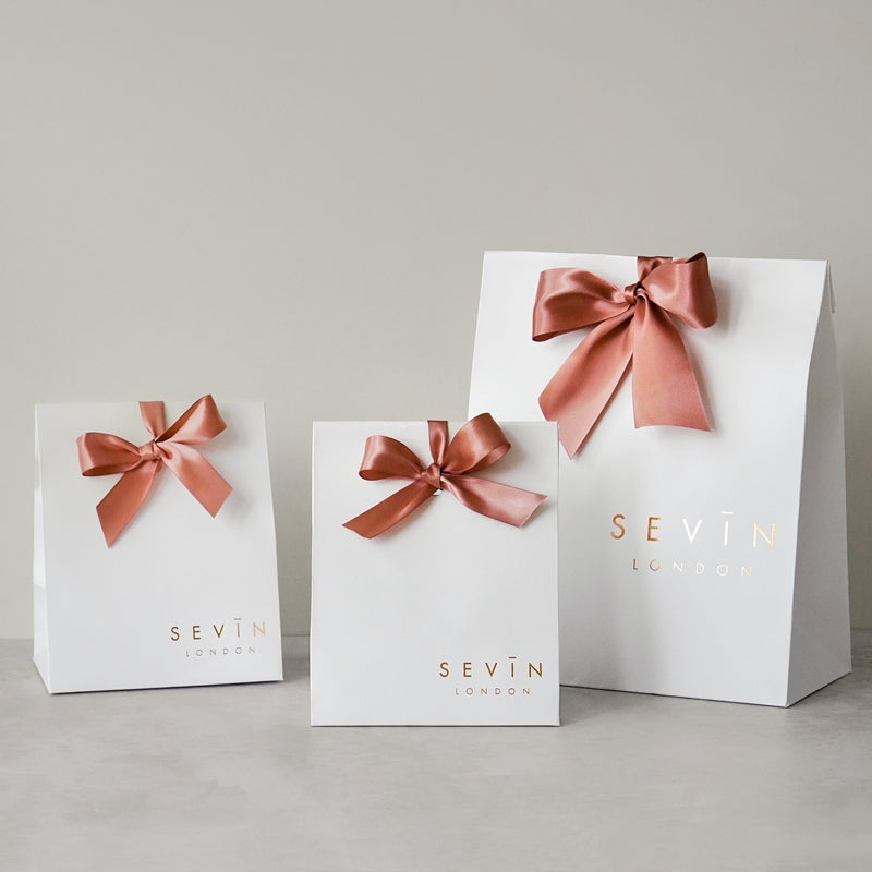 Three white gift bags with pink ribbons and 'Sevin London' branding on a light gray background.