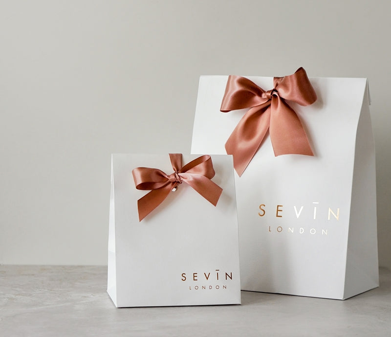 Two white gift bags with bronze ribbons and 'Sevin London' branding on a light gray background.