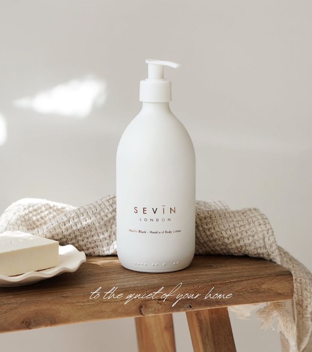 A matte white pump bottle of SEVIN LONDON Hand and Body Wash stands on a rustic wooden stool, styled with a beige knit throw blanket and a bar of soap on a small white dish.